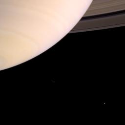 Saturn's atmosphere is prominently shown with the rings emerging from behind the planet at upper right. The two moons on the left of the image, Mimas and Enceladus, were captured by NASA's Cassini spacecraft.