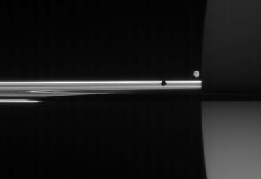 The unlit side of the rings glows with scattered sunlight as two moons circle giant Saturn. The light reaching NASA's Cassini spacecraft in this view has traveled many paths before being captured.