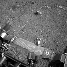 This image shows a close-up of track marks from the first test drive of NASA's Curiosity rover. The rover's arm is visible in the foreground. A close inspection of the tracks reveals a unique, repeating pattern: Morse code for JPL.