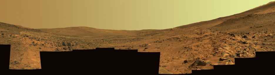 This panorama shows two reddish-brown, rock-strewn slopes on the left and right sides of a broad, U-shaped dip in the middle. In the distance are the broad slopes of 'McCool Hill.' Above that is an orangish-yellow sky.