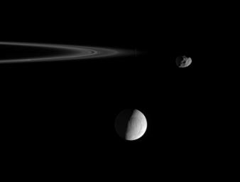 Wrinkled and cracked Enceladus hangs in the distance as the pitted ring moon Janus, at right, rounds the outer edge of the F ring. This image was taken in visible light with NASA's Cassini spacecraft's narrow-angle camera.