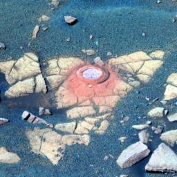 This false-color image shows a circular indentation in a flat-topped rock surface. Around the edge of the hole is a fine layer of reddish dust. The rock is light tan with a moderately cracked the surface. Around it is a layer of bluish sand and pebbles.