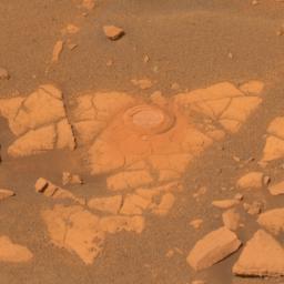 This true-color image shows a circular indentation in a flat-topped rock surface. Around the edge of the hole is a fine layer of dust. The rock has a moderately cracked the surface. Around it is a layer of sand and pebbles. The view is reddish brown.