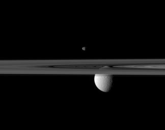 Saturn's rings occupy the foreground of this image captured by NASA's Cassini spacecraft. The small moon Janus appears to hover above, while the far larger moon Rhea is partially obscured by the rings.