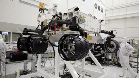 This image was taken in the cleanroom where NASA's Curiosity rover is being assembled. It shows the rover, which is about the size of an SUV, hoisted on a white lift, with its black wheels suspended in the air.