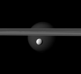 The brightly reflective moon Enceladus appears before Saturn's rings while the larger moon Titan looms in the distance. Jets of water ice and vapor emanating from the south pole of Enceladus (hinting at subsurface sea rich in organics).