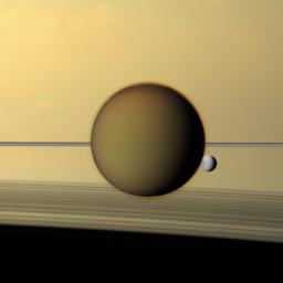 Saturn's fourth-largest moon, Dione, can be seen through the haze of the planet's largest moon, Titan, in this view of the two posing before the planet and its rings from NASA's Cassini spacecraft.