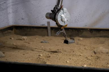 The 3D-printed titanium scoop of the Cold Operable Lunar Deployable Arm (COLDArm) robotic arm system is poised above a test bed filled with material to simulate lunar regolith (broken rocks and dust) at NASA's Jet Propulsion Laboratory.