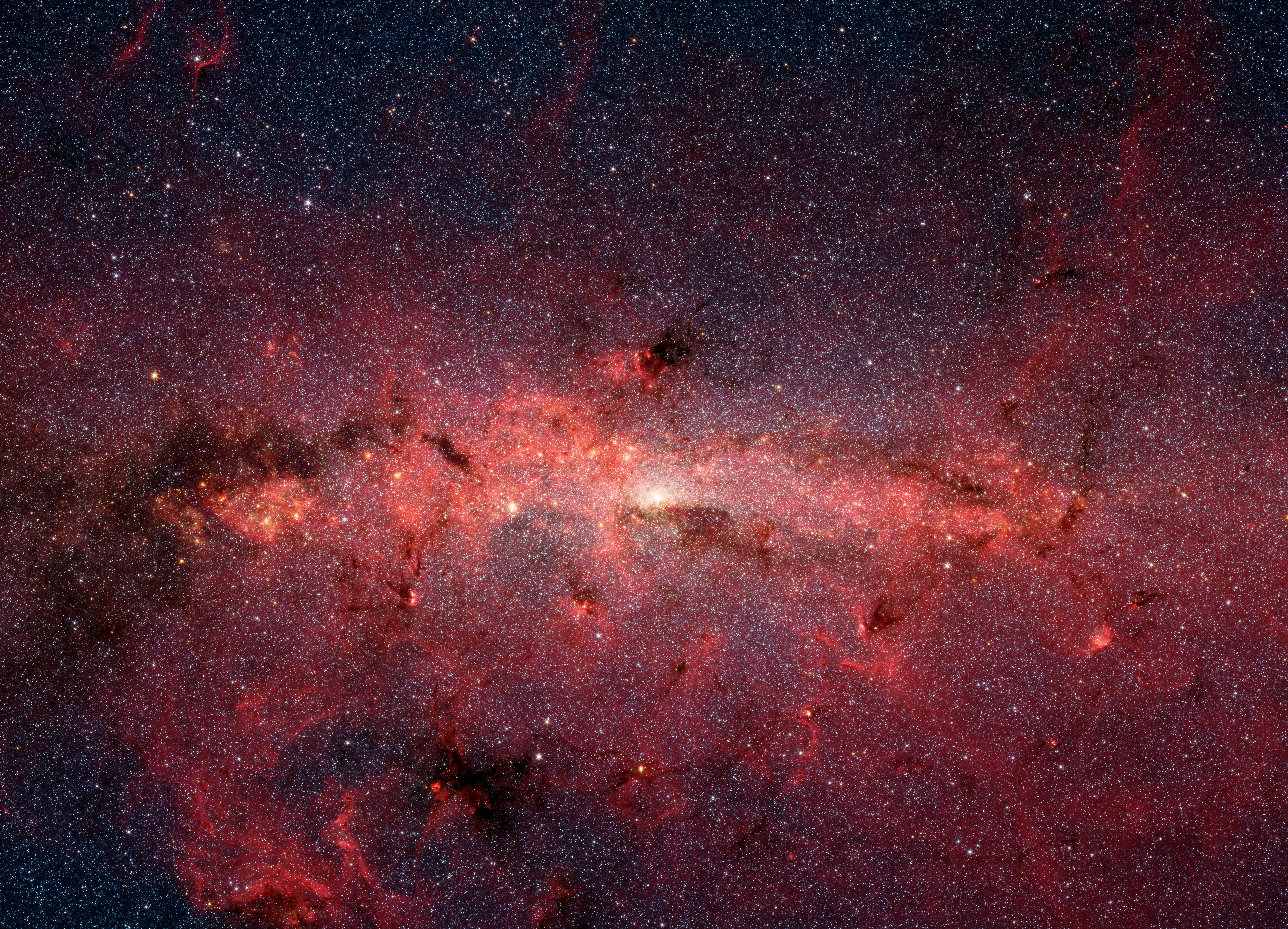 A bright band of crimson-colored dust stretches across the center of this image covered in tiny specs of light from hundreds of thousands of stars.